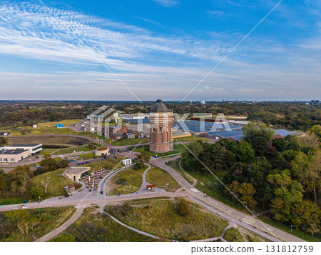 Aerial view of historical water tower amid utility buildings, green paths, and distant city skyline blending heritage architecture with infrastructure and natural surroundings. Aerial view of historical water tower amid utility buildings, green paths, and distant city skyline blending heritage architecture with infrastructure and natural surroundings. 131812759