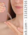 Young woman holding a dropper with serum for face. Vertical macro view pink background. Dermatology, cosmetics, spa ads. Liquid skincare product, beauty concept. Facial oil fall from a glass pipette  131812916