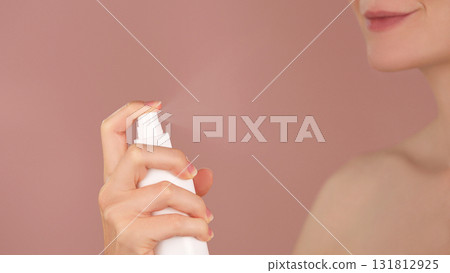 Skincare ads. Young woman sprays a facial spray mist or thermal water, face toner, facial tonic. Spraying process. Toning, refreshing, moisturizing, soothing skin. Pink background. Copy space  131812925