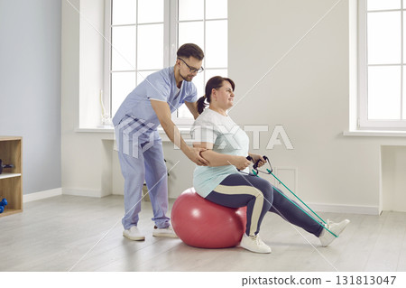 Nurse man helping fat patient woman to do stretching exercises with rubber band sitting on fit ball 131813047
