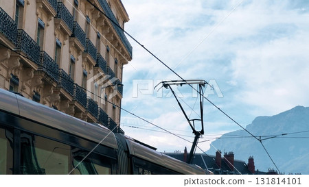 Close-up view of a modern tram passing through Grenoble, France. Historic building facades, power lines, and alpine mountains visible in the background 131814101