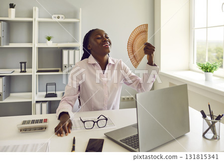 African American woman office worker refreshes herself with hand fan at work in hot summer African American woman office worker refreshes herself with hand fan at work in hot summer 131815341