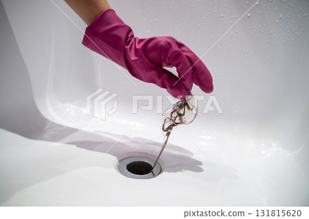 Hand of woman in pink rubber glove taking off hair clump from water drain, blocked pipe in bath. 131815620