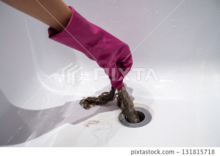 Hand of woman in pink rubber glove taking off hair clump from water drain, blocked pipe in bath. 131815718