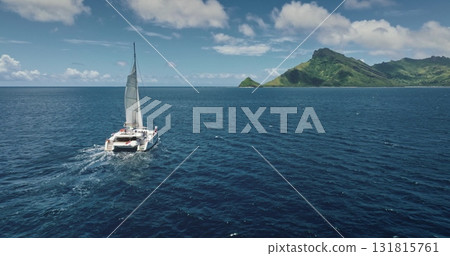 Fiji, Kuata island: White catamaran with tourists sailing blue ocean water near tropical island, under bright sunny sky. Remote wild nature paradise, exotic summer travel. Aerial drone flight 131815761