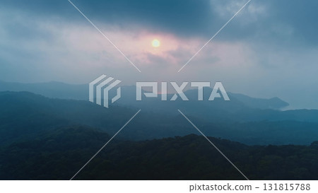 Dark green forested hills and mountains fading into the misty distance with sun setting behind clouds illuminating sky with soft orange and pink light 131815788