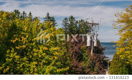 Electric utility pole with multiple transformers surrounded by colorful autumn trees and overlooking the coast in the United States, symbolizing modern electrification and energy distribution. 131818223