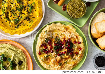 A variety of hummus flavors including turmeric, classic, and broccoli are served with fresh bread, nuts, and garnishes on a colorful charcuterie board, perfect for sharing 131820380