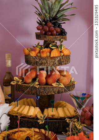 Festive Fruit Tiered Stand with Pineapple, Grapes, Peaches, Cheese, and Wine for a Celebration Event, Rustic Wood and Pink Backdrop. Festive Fruit Tiered Stand with Pineapple, Grapes, Peaches, Cheese, and Wine for a Celebration Event, Rustic Wood and Pink Backdrop. 131829495