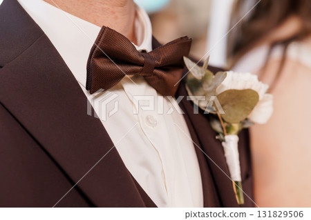 Elegant Wedding Groom Detail: Brown Bow Tie and Boutonniere with White Roses on Black Tuxedo, Close-Up View, Romantic Celebration. Elegant Wedding Groom Detail: Brown Bow Tie and Boutonniere with White Roses on Black Tuxedo, Close-Up View, Romantic Celebration. 131829506