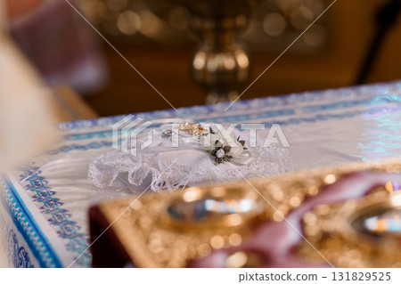 Intimate Wedding Ring Ceremony Detail: Gold Bands on White Pillow with Lace, Floral Accent in Church Setting. Intimate Wedding Ring Ceremony Detail: Gold Bands on White Pillow with Lace, Floral Accent in Church Setting. 131829525