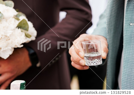 Groom Holding Shot Glass at Wedding, Bouquet and Suit Details, Soft Focus, Brown and Gray Formal Attire, Celebration Tradition. Groom Holding Shot Glass at Wedding, Bouquet and Suit Details, Soft Focus, Brown and Gray Formal Attire, Celebration Tradition. 131829528