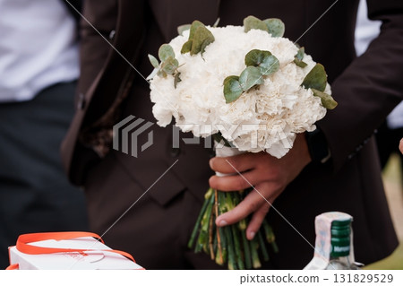 Elegant Wedding Boutonniere Held by Groom's Hands, White Carnations, Eucalyptus, Brown Suit, Celebration, Formal Event Photography. Elegant Wedding Boutonniere Held by Groom's Hands, White Carnations, Eucalyptus, Brown Suit, Celebration, Formal Event Photography. 131829529