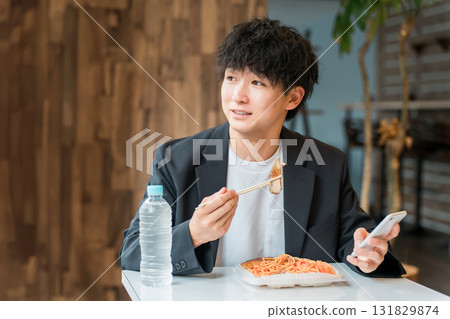 一位年輕的亞洲男性商人一邊吃盒飯,一邊在餐廳看著智慧型手機 一位年輕的亞洲男性商人一邊吃盒飯,一邊在餐廳看著智慧型手機 131829874