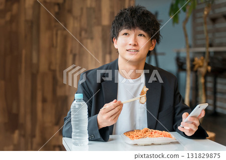 一位年輕的亞洲男性商人一邊吃盒飯,一邊在餐廳看著智慧型手機 一位年輕的亞洲男性商人一邊吃盒飯,一邊在餐廳看著智慧型手機 131829875
