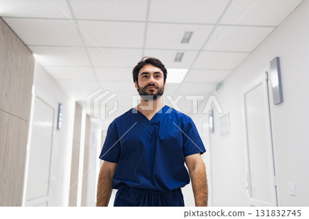 A doctor in blue scrubs walks down a bright hallway in a clinic or hospital setting. A doctor in blue scrubs walks down a bright hallway in a clinic or hospital setting. 131832745