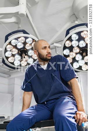 A surgeon in blue scrubs sits on an operating table, looking thoughtful under bright surgical lights. A surgeon in blue scrubs sits on an operating table, looking thoughtful under bright surgical lights. 131832758