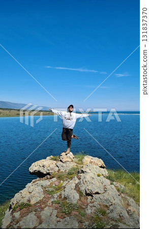 Person posing triumphantly on a rocky cliff overlooking a vast blue lake Person posing triumphantly on a rocky cliff overlooking a vast blue lake 131837370