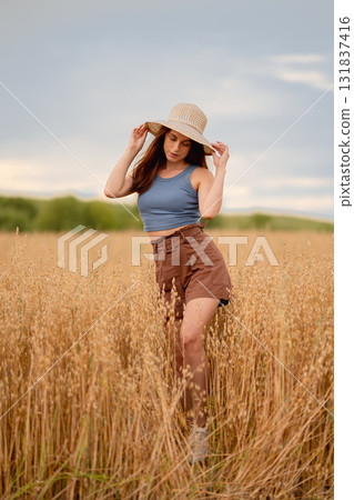 Young woman wearing a hat poses in an oat and wheat field during golden hour Young woman wearing a hat poses in an oat and wheat field during golden hour 131837416