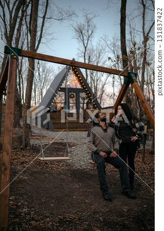 Couple enjoys a quiet moment on a swing in front of a cozy cabin in the woods during dusk 131837422