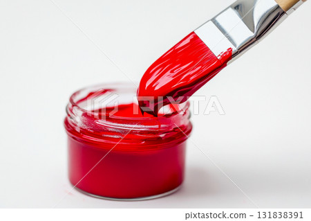 A brush dipped in bright red paint is poised above a small jar, showcasing the vibrant color against a white background 131838391