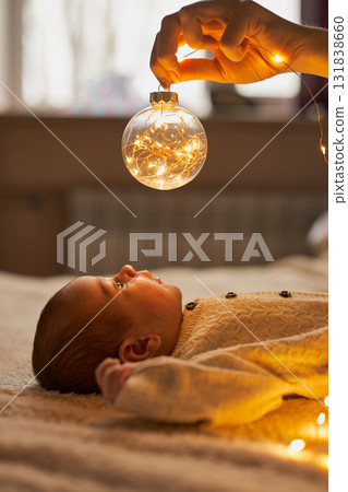 Newborn baby in a knit romper lies on a cozy blanket, gazing up in wonder at a clear Christmas 131838660