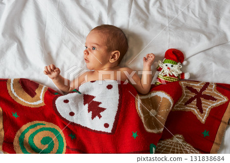 An awake newborn baby lies on a white sheet, partially covered by a cozy, red Christmas blanket with An awake newborn baby lies on a white sheet, partially covered by a cozy, red Christmas blanket with 131838684