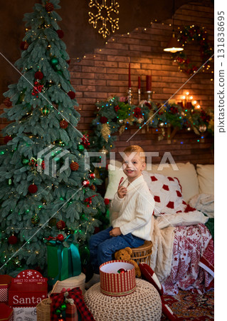 A young boy in a white sweater sits next to a Christmas tree and reaches out to carefully hang a red A young boy in a white sweater sits next to a Christmas tree and reaches out to carefully hang a red 131838695