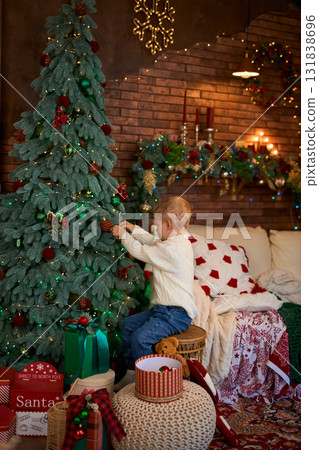 A young boy in a white sweater sits next to a Christmas tree and reaches out to carefully hang a red A young boy in a white sweater sits next to a Christmas tree and reaches out to carefully hang a red 131838696