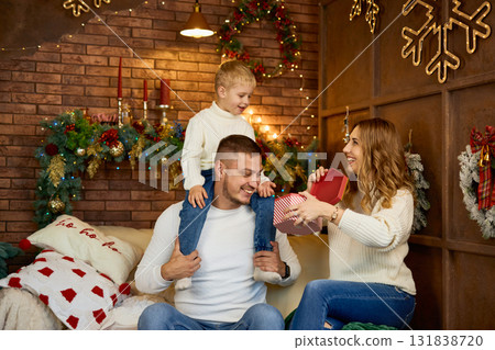 Happy family in white sweaters exchanging a Christmas gift in a cozy loft-style room with a brick 131838720