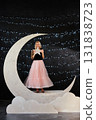 A blonde woman in a black top and a voluminous pink tulle skirt poses on a large, glitter crescent 131838723