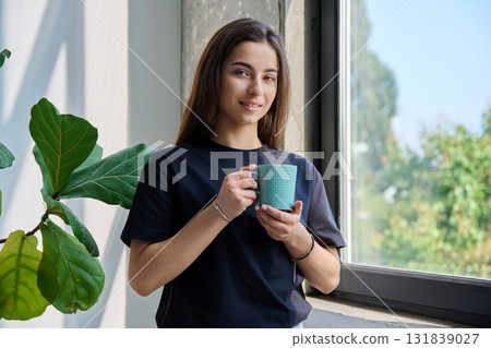 Portrait of smiling relaxed teenage girl posing with cup near window 131839027