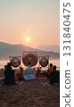 Participants engage in a sound therapy session at sunrise on the beach, surrounded by calming gongs and the serene backdrop of mountains reflecting soft morning light 131840475