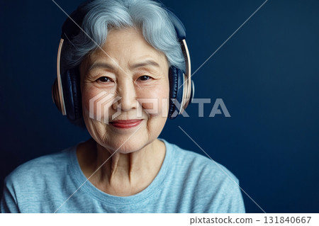 A smiling Asian senior woman with headphones on her head, set against a dark blue background, embracing the pleasure of listening to music or studying, radiating the joy of life in old age 131840667