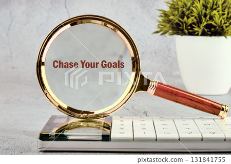 A magnifying glass rests on a keyboard, highlighting the text Chase Your Goals. A potted plant is in the background, signifying growth and achievement in business 131841755