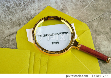 A golden magnifying glass focuses on a paper labeled Entrepreneurship 2026 resting on a stack of bright yellow envelopes, representing planning and strategic thinking for future success 131841756