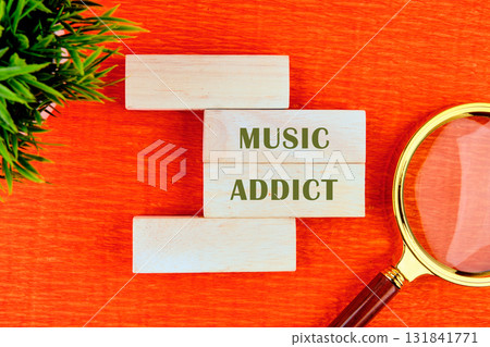 This setup has white wooden blocks with green letters spelling out MUSIC ADDICT. A magnifying glass sits beside them, and a small potted plant is in the corner of an orange surface This setup has white wooden blocks with green letters spelling out MUSIC ADDICT. A magnifying glass sits beside them, and a small potted plant is in the corner of an orange surface 131841771