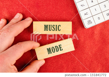 A hand carefully positions wooden blocks displaying the words MUSIC MODE on a bright red background, next to a white calculator A hand carefully positions wooden blocks displaying the words MUSIC MODE on a bright red background, next to a white calculator 131841772