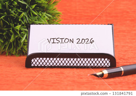 A card reading Vision 2026 sits on a desk, accompanied by a fountain pen and a small green bush. The setting suggests planning for the future A card reading Vision 2026 sits on a desk, accompanied by a fountain pen and a small green bush. The setting suggests planning for the future 131841791