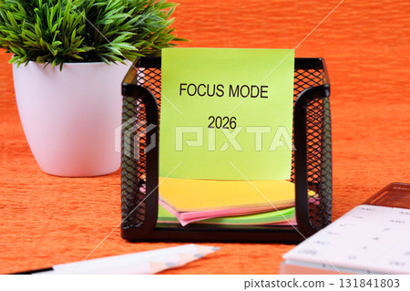 A green sticky note labeled Focus Mode 2026 sits in a black wire holder on an orange desk. A potted plant, pen, and calculator are nearby 131841803