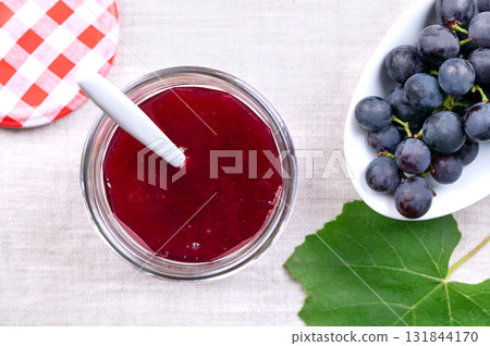 Grape jam made of blue grapes, in glass jar on linen. Open glass with screw cap and plastic spoon dipped in homemade grape jam. On the right fresh leaf and grapes of common grape vine, Vitis vinifera. Grape jam made of blue grapes, in glass jar on linen. Open glass with screw cap and plastic spoon dipped in homemade grape jam. On the right fresh leaf and grapes of common grape vine, Vitis vinifera. 131844170