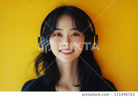 A smiling Asian girl with headphones on her head, set against a yellow background, embodying the pleasure of listening to music or studying in a relaxed and joyful moment 131846783