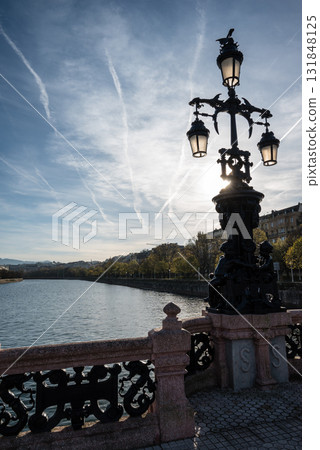 Street lights on a large historic bridge bathed in the setting sun under a sky full of contrails in San Sebastian 131848125