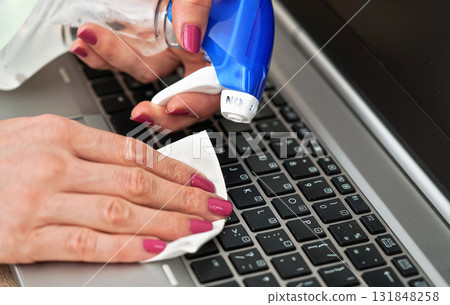 Woman cleaning laptop keyboard with white tissue, detail on her fingers holding paper towel, blue alcohol sprayer near - disinfection concept 131848258