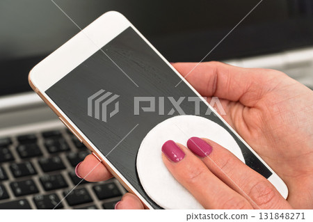 Woman cleaning mobile phone with cotton paper pad, wiping black screen, closeup detail. Blurred laptop keyboard in background Woman cleaning mobile phone with cotton paper pad, wiping black screen, closeup detail. Blurred laptop keyboard in background 131848271