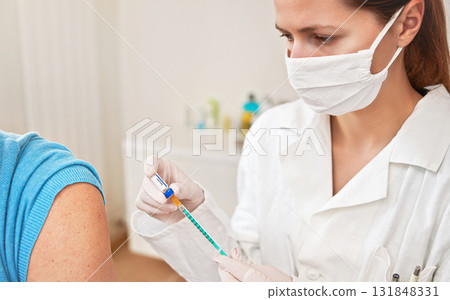 Female doctor in white coat and cotton face mask injecting vaccine into syringe, about to inject it to patient shoulder near Female doctor in white coat and cotton face mask injecting vaccine into syringe, about to inject it to patient shoulder near 131848331