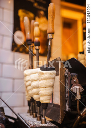 Traditional Czech chimney cakes called Trdelnik baking on rotating metal rods in a Prague street bakery, showing the preparation process of this famous sweet pastry. 131848994