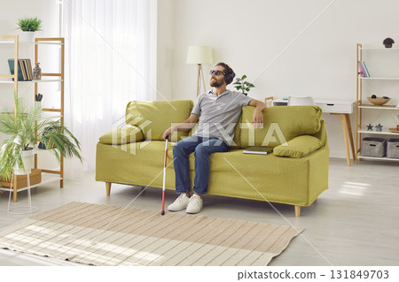 Young smiling blind man sitting on sofa at home in headphones and listening to music or audiobook. Young smiling blind man sitting on sofa at home in headphones and listening to music or audiobook. 131849703