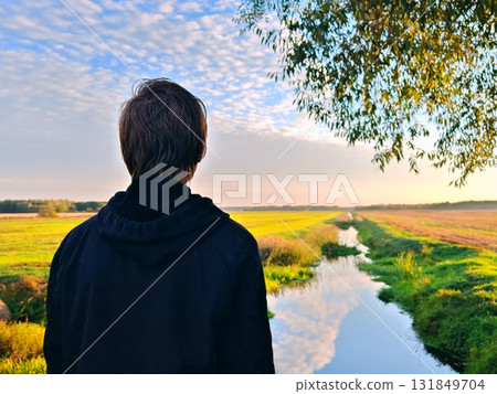 Guy is standing with his back to camera in an autumn landscape in countryside. A man in a hoodie stands with his back to the camera against a beautiful forest lake. Enjoy nature and travel 131849704