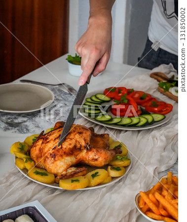 Medium view of a hand cutting roasted chicken at a festive holiday table decorated with salads, mandarins, and sweets. Cozy Christmas and New Year celebration with joyful atmosphere. 131850002